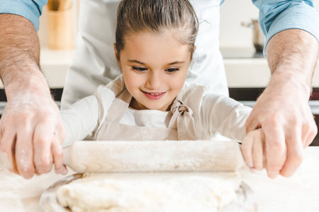 young smiling kid with her father rolling out dough at kitchen