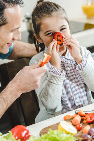 happy father with daughter, little kid applies pepper slice to nose and looking at camera at kitchenの写真素材