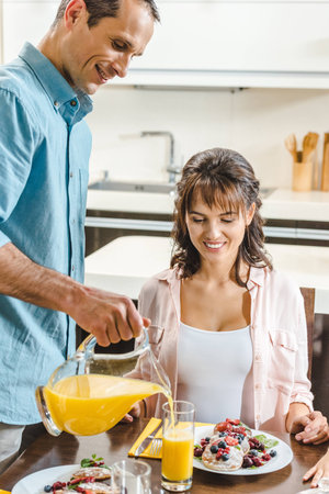 happy couple, man pouring juice in glass for sitting at table pregnant woman with pancakes at...