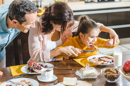 cheerful family together eating pancakes with berries at table, mother feeding daughterの写真素材