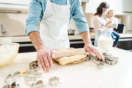 cropped image of man rolling out dough by rolling pin on table in front of mother with daughter at...