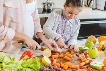 partial view of mother with daughter slicing carrots on chopping board at kitchenの写真素材