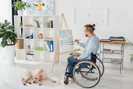 disabled man on wheelchair painting on canvas while his dog laying on floorの写真素材