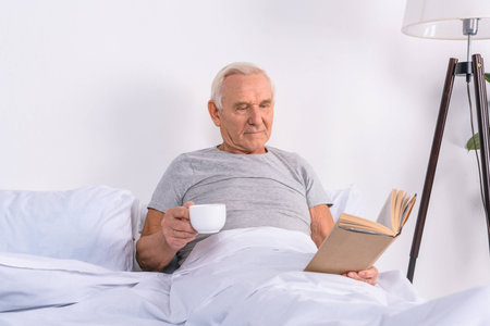 portrait of senior man with cup of coffee reading book in bed at homeの写真素材