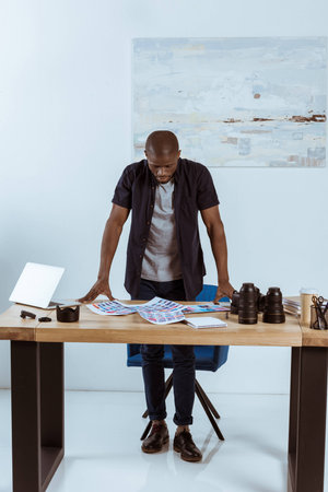 concentrated african american photographer looking at photoshoot examples while standing at workplaceの写真素材
