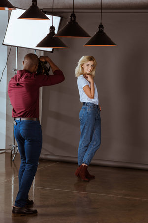 beautiful smiling caucasian woman posing while african american photographer taking picture in studioの写真素材