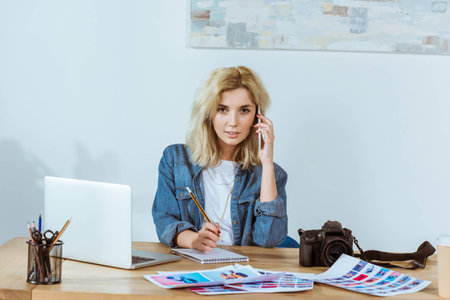 portrait of young photographer talking on smartphone while sitting at workplace in studioの写真素材
