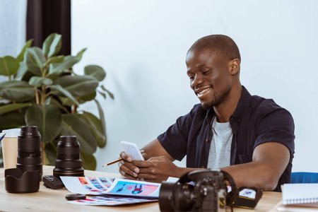 portrait of smiling african american man using smartphone while sitting at workplace with...