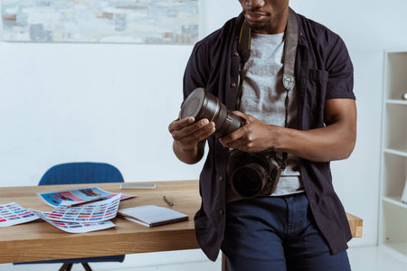 partial view of african american photographer with photo camera leaning on workplace in officeの写真素材
