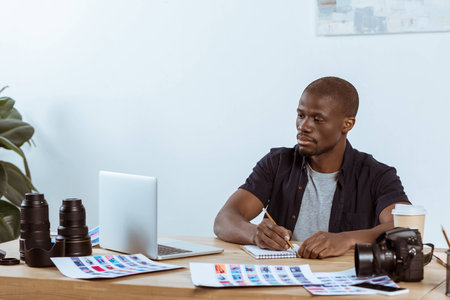 portrait of concentrated african american photographer working at workplace with laptop in officeの写真素材