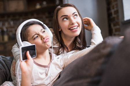 portrait of grimace daughter listening music in headphones with smiling mother resting on sofa near byの写真素材