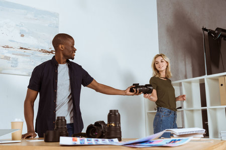 smiling caucasian photographer giving photo camera to african american colleague at workplace in studioの写真素材