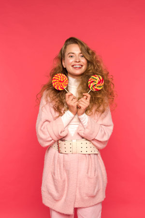 happy stylish girl standing with two lollipops and looking at camera isolated on redの写真素材