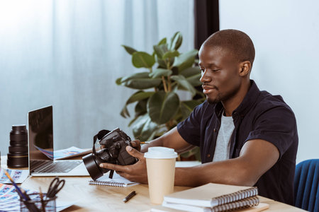 portrait of african american photographer with photo camera at workplace with laptop in officeの写真素材