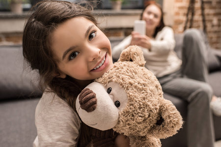 selective focus of smiling daughter with teddy bear and mother using smartphone at homeの写真素材