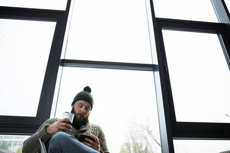 bottom view of man holding thermal cup and looking at smartphone