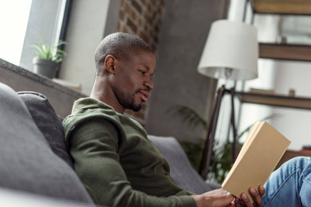 side view of young african american man reading book at homeの写真素材