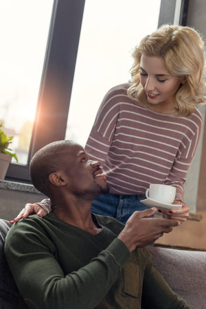 portrait of woman brought cup of coffee to smiling african american boyfriendの写真素材