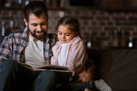 happy father with daughter hugging on couch and reading book at homeの写真素材