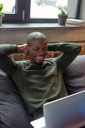 portrait of smiling african american man with laptop resting on sofa at homeの写真素材