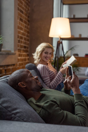 selective focus of african american man using smartphone while caucasian girlfriend reading book on sofa at homeの写真素材