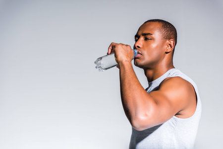 young african american sportsman drinking water from bottle isolated on greyの写真素材