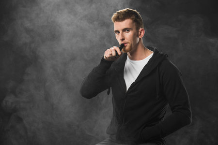Young bearded man vaping electronic cigarette surrounded by clouds of steam