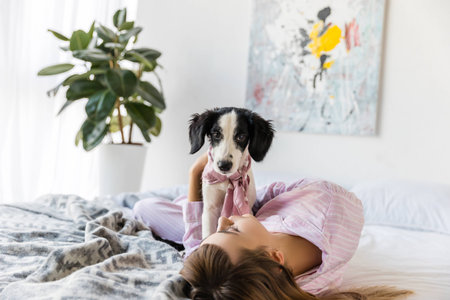 young woman in pajamas lying on bed together with black and white puppyの写真素材