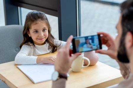 cropped image of father taking photo of smiling daughter in cafe