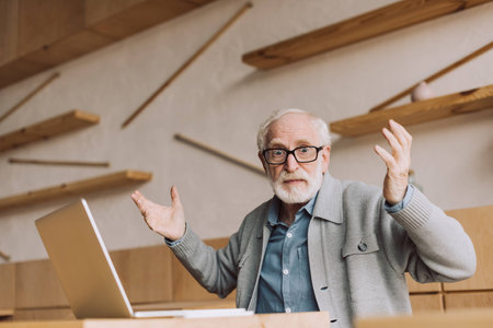 bewildered senior man with laptop sitting in cafe and looking at camera with raised hands