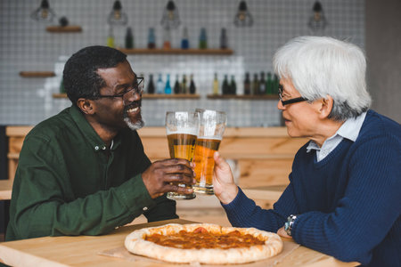 smiling senior friends clinking glasses of beer at bar with pizza on table