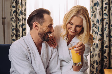 happy middle aged couple in bathrobes drinking juice during breakfast in hotel roomの写真素材