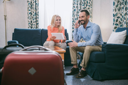 smiling mature couple using smartphone and digital tablet while sitting with suitcases in hotel roomの写真素材