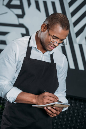 handsome young african american waiter writing in notepadの写真素材