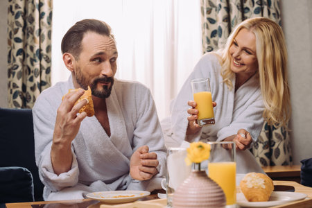smiling middle aged couple drinking juice and eating pastry during breakfast in hotel roomの写真素材