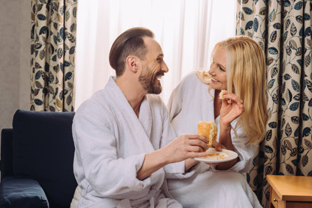 happy mature couple in bathrobes eating pastry for breakfast together in hotel roomの写真素材