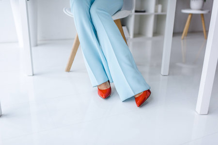 cropped view of woman in trendy flared jeans and red heels sitting at tableの写真素材