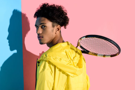Young bright african american guy holding tennis racket on shoulder on pink and blue backgroundの写真素材