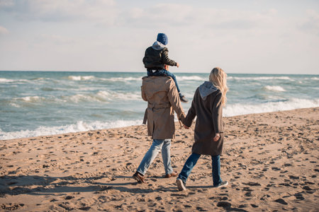 back view of parents with son on shoulders walking on seashore in autumnの写真素材