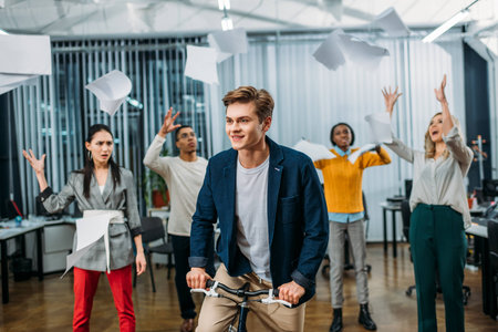 young businessman riding bike at office while partners throwing up papers on background