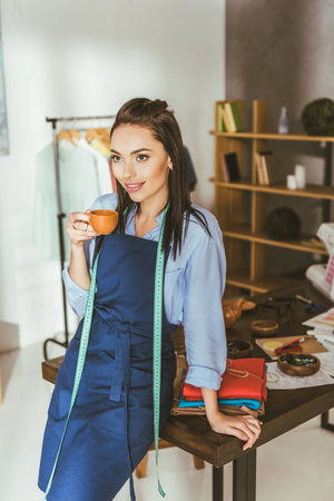 attractive seamstress leaning on table, holding cup of tea and looking awayの写真素材