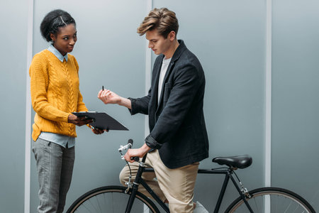 handsome young businessman signing contract in hands of young african american colleague while sitting on bikeの写真素材