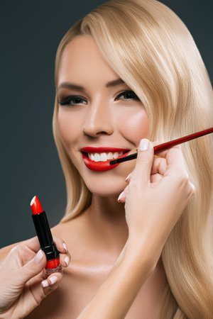 Portrait of beautiful blonde hair girl with visagiste hands applying lipstick with brush, isolated on blackの写真素材