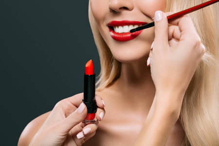 cropped image of beautiful blonde hair girl with visagiste applying lipstick with brush, isolated on blackの写真素材