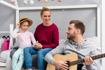 happy young father playing guitar for wife and daughter at kid bedroom