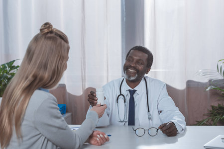 Smiling african american doctor giving plastic jar with pills to patientの写真素材