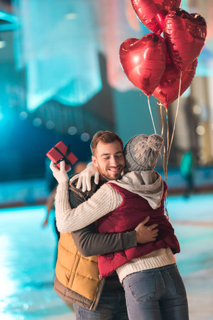 young woman holding gift box and hugging happy boyfriend with heart shaped balloons on rinkの写真素材