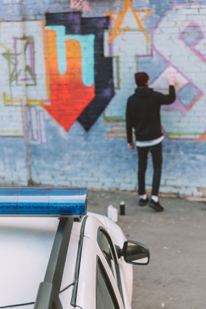 back view of vandal painting graffiti on wall, police car on foregroundの写真素材