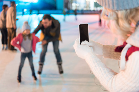 cropped shot of woman holding smartphone with blank screen and photographing family skating on rinkの写真素材