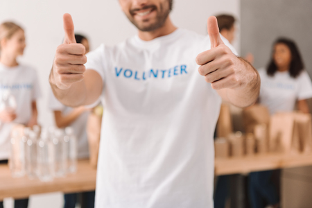 cropped shot of handsome volunteer showing thumbs up with blurred colleagues working on backgroundの写真素材